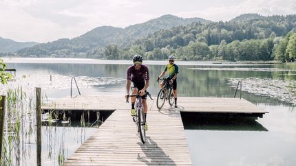 Gravel-Wundertüte Wörthersee © Region Wörthersee Rosental Tourismus GmbH - Chris Perkles Zwei Radfahrer fahren auf einem Steg über einen ruhigen See mit Bergen im Hintergrund