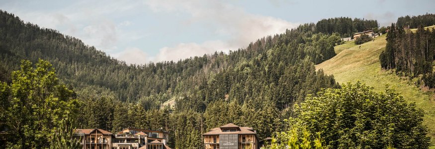 Prädikat: Außergewöhnlich © Hannes Niederkofler Hotelgebäude eingebettet in grüne Berge und Wald unter blauem Himmel