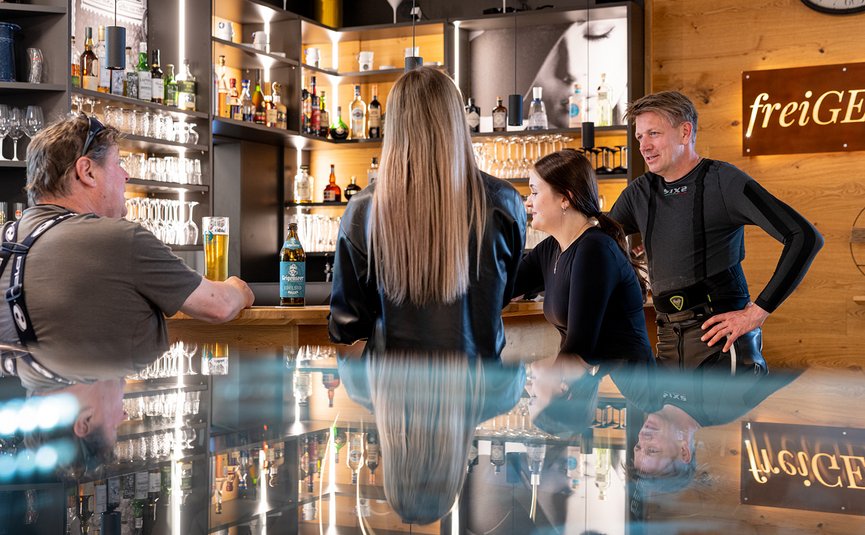DAS UNTERRAIN lifestyle hotel **** © Armin Kleinlercher Menschen unterhalten sich an einer Bar mit Getränken und Holzinterieur
