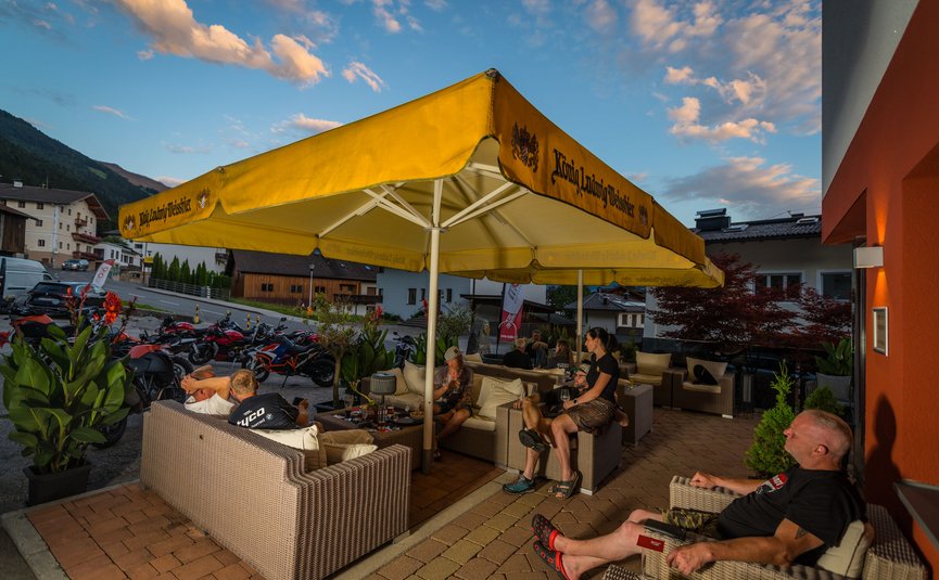 Personen sitzen unter gelbem Sonnenschirm auf Terrasse mit Motorrädern im Hintergrund