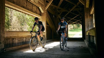 into the wold © Bregenzerwald Tourismus - Martin Granadia Zwei Radfahrer fahren durch eine hölzerne gedeckte Brücke bei Tageslicht