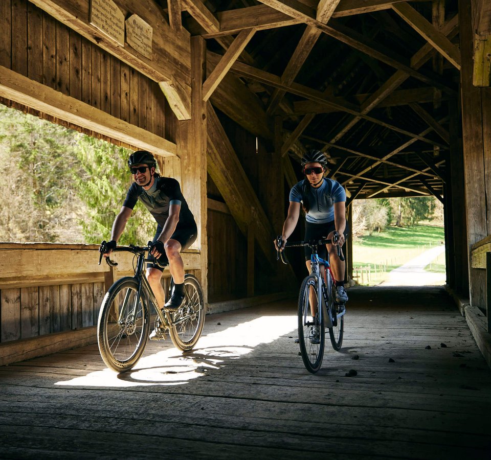 into the wold © Bregenzerwald Tourismus - Martin Granadia Zwei Radfahrer fahren durch eine hölzerne gedeckte Brücke bei Tageslicht