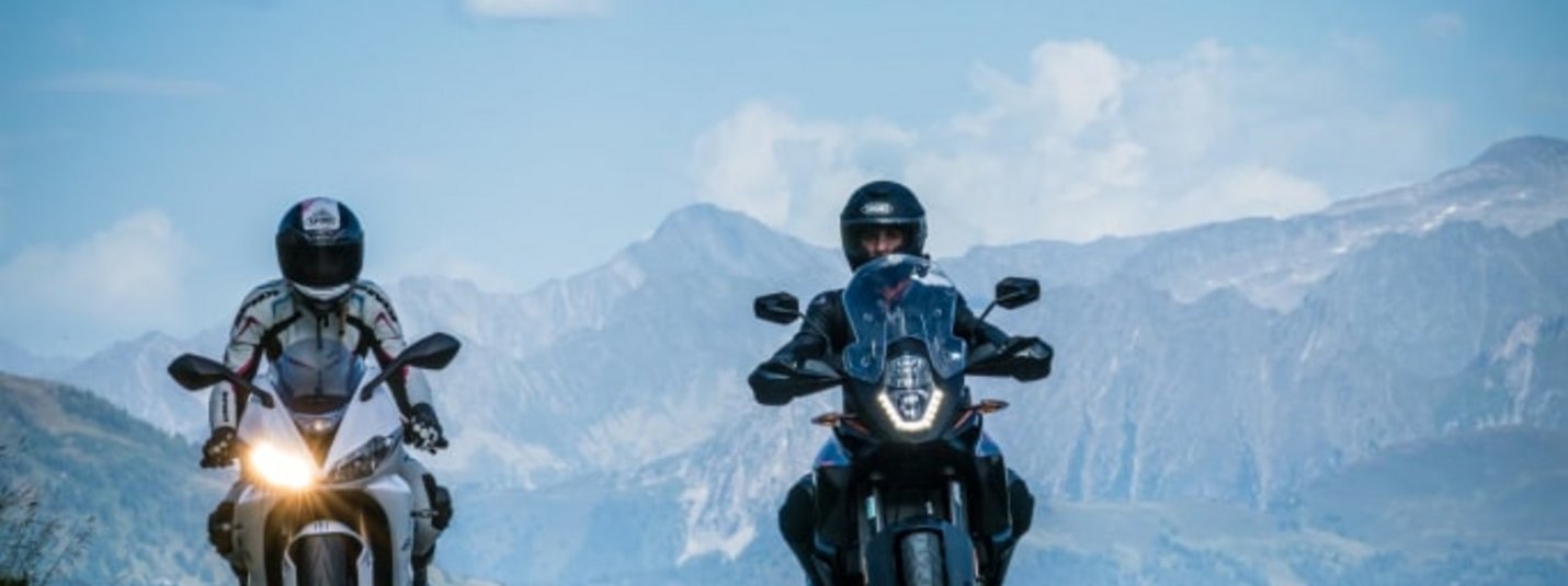 5 Tages Motorrad Angebot Zwei Motorradfahrer fahren vor Bergen unter einem bewölkten Himmel