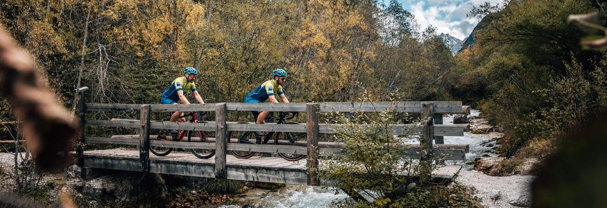 Gravel-Wochenende in San Vigilio © Jens Scheibe Zwei Radfahrer überqueren eine Holzbrücke über einen Bach im Wald