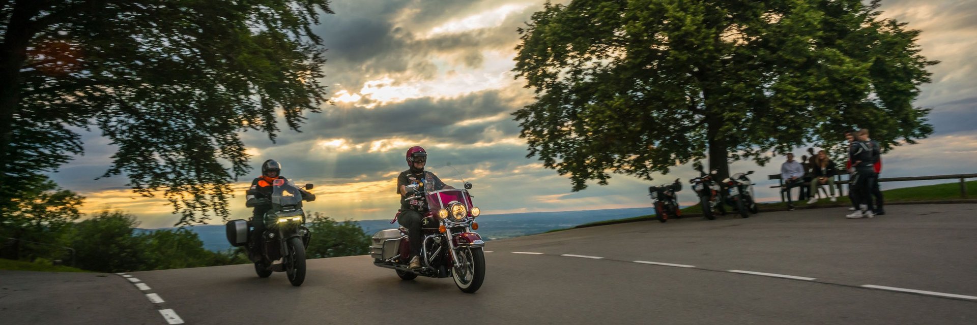 Allgäu © Moppetfoto.de Zwei Motorradfahrer fahren bei Sonnenuntergang an einer Kurve vorbei.