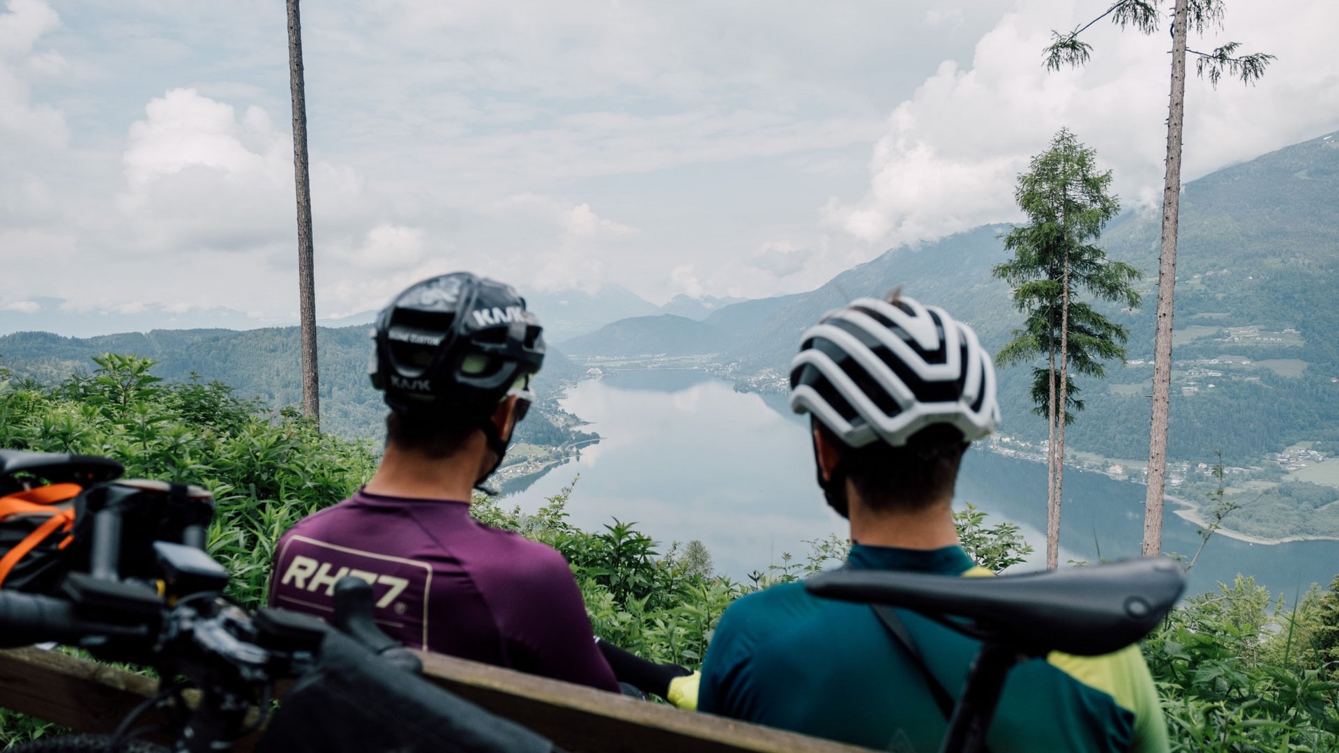 Gravel-Wundertüte Wörthersee © Region Wörthersee Rosental Tourismus GmbH - Chris Perkles Zwei Radfahrer mit Helmen sitzen auf einer Bank und schauen auf einen See in den Bergen