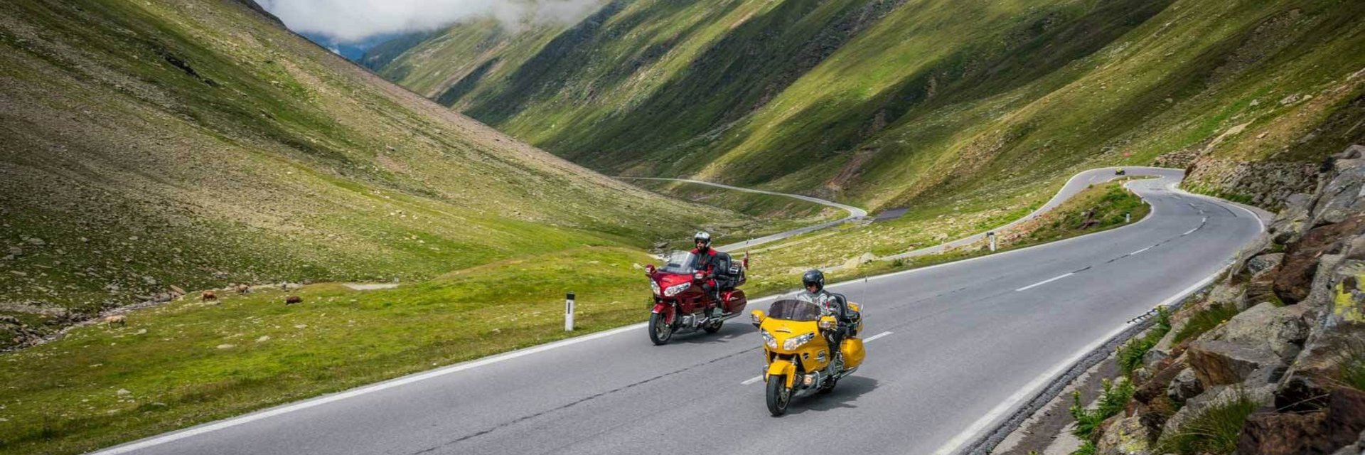 Zwei Motorräder fahren auf einer Bergstraße mit grünen Hügeln und Wolken