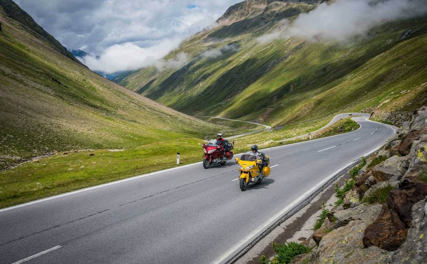 Motorradurlaub am Arlberg – Dreiländereck © Moppetfoto.de Zwei Motorräder fahren auf einer Bergstraße mit grünen Hügeln und Wolken