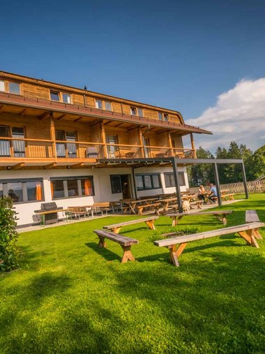 Motorradurlaub © Moppetfoto.de Holzhaus mit Terrasse und Sitzbänken auf grünem Rasen unter blauem Himmel