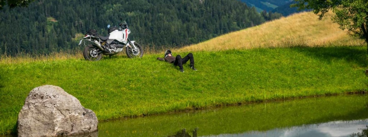 Biken ohne Stress Person liegt auf grüner Wiese mit Motorrad neben Teich und Bergkulisse