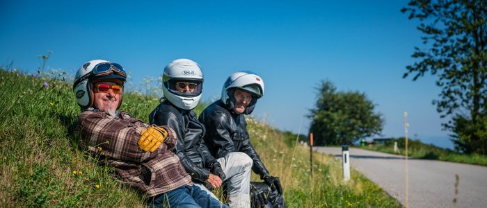 Waldviertel © Moppetfoto.de Drei Motorradfahrer in Helmen sitzen auf einer Wiese neben einer Straße.