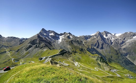 Motorradurlaub: Großglockner – Hohe Tauern © Großglockner Hochalpenstraßen AG Serpentinenstraße in grünen Alpen mit schneebedeckten Bergen unter blauem Himmel
