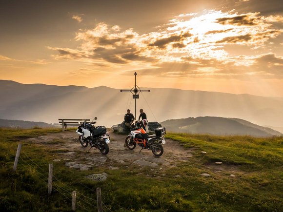 Zwei Motorräder und zwei Personen bei einem Gipfelkreuz im Sonnenuntergang in den Bergen