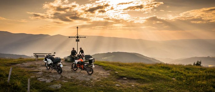 Zwei Motorräder und zwei Personen bei einem Gipfelkreuz im Sonnenuntergang in den Bergen