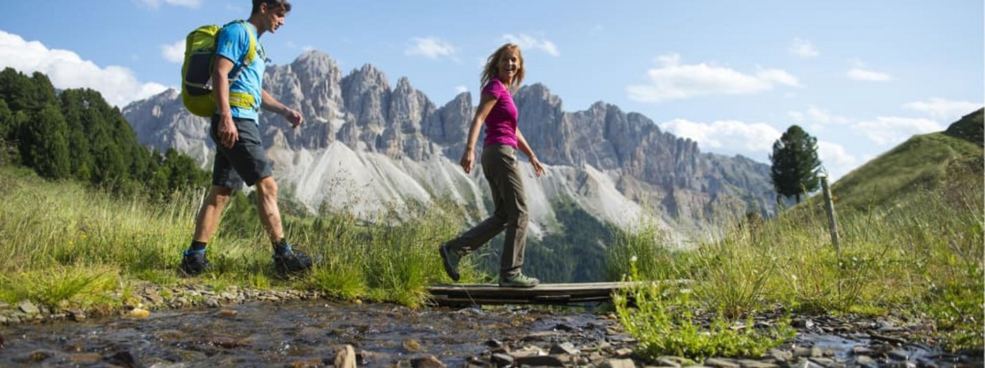 Berge und Natur Wanderwochen Zwei Menschen wandern über eine Holzbrücke an einem Bergbach mit Alpen im Hintergrund