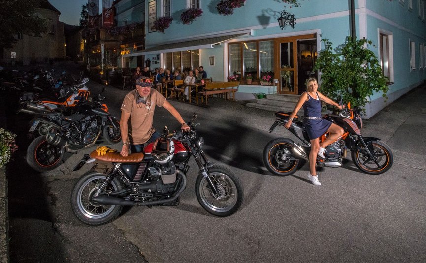 Zwei Personen mit Motorrädern vor blauem Haus bei Nacht
