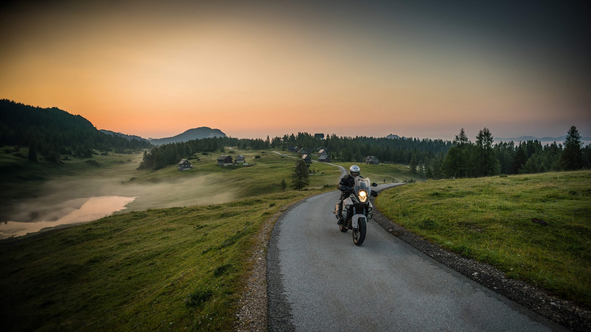 Motorradfahrer fährt auf Landstraße bei Sonnenuntergang mit Hütten und Bergen