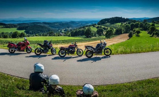 Biker Week Drei Personen mit Helmen sitzen auf einer Wiese vor vier Motorrädern auf einer Landstraße