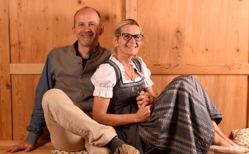 Hotel & Südtiroler Gasthaus Der Gassenwirt *** © Hotel Gassenwirt Lächelndes Paar in traditioneller Tracht sitzt auf Holzbank vor Holzwand