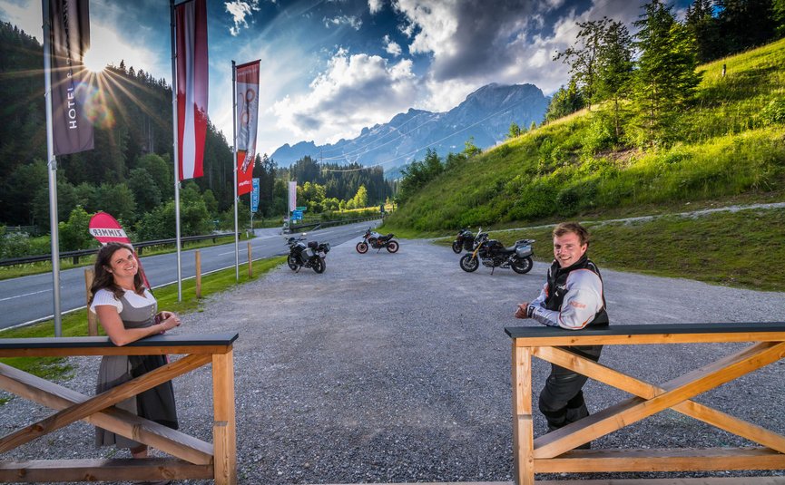 Mann und Frau an Holzzaun vor Motorrädern und Bergen bei sonnigem Himmel