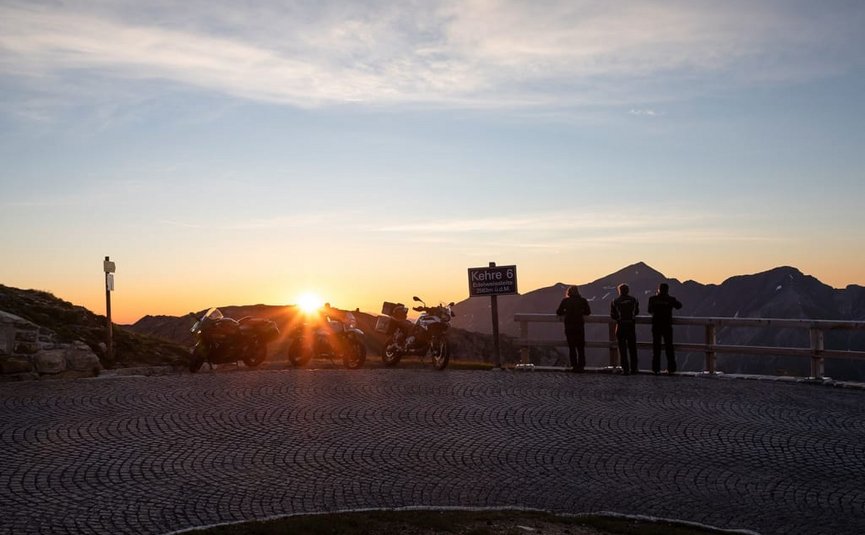 Sternfahrt Motorrad-Special ab € 598,- pro Person Motorräder und Menschen bei Sonnenaufgang an einem Bergpass mit Schild Kehre 6