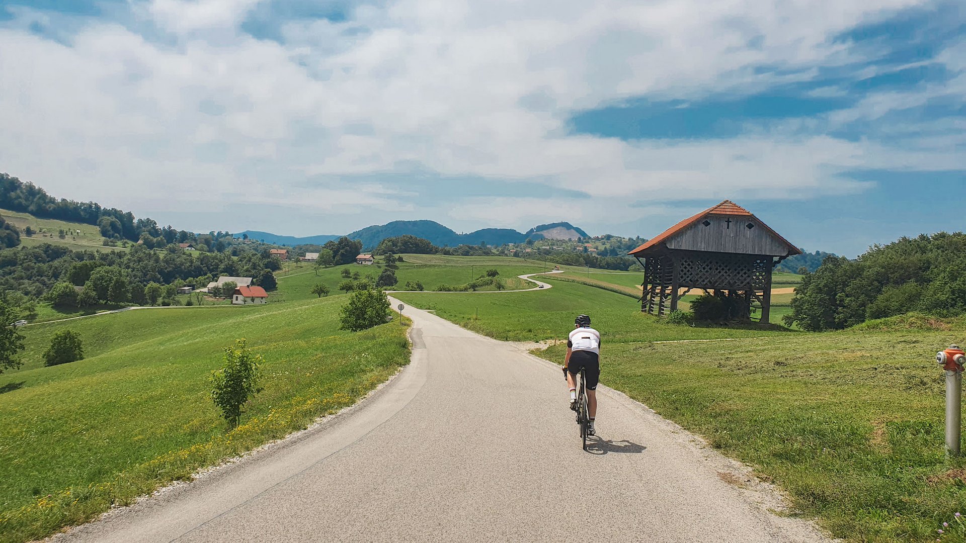 Radfahrer auf Landstraße mit grünem Hügel und traditionellem Holzbau im Hintergrund