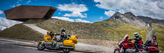 Motorradurlaub am Arlberg – Dreiländereck © Moppetfoto.de Zwei Motorradfahrer auf Bergstraße mit Bergen und modernem Gebäude im Hintergrund