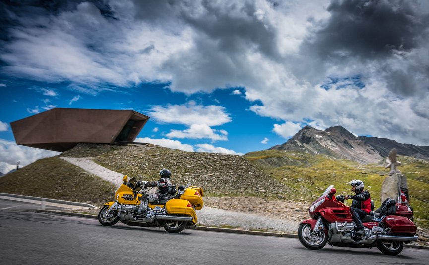 Hotel Jägerhof **** © Moppetfoto.de Zwei Motorradfahrer auf Bergstraße mit Bergen und modernem Gebäude im Hintergrund