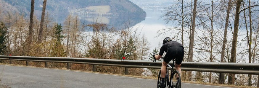 Gravelabenteuer zwischen See & Nockbergen © Martin Hoffmann Radfahrer fährt bergab auf asphaltierter Straße mit See und Bergen im Hintergrund