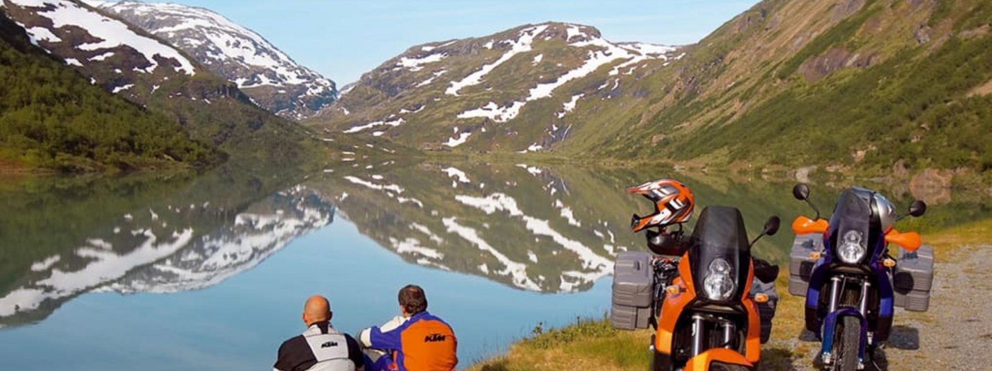 Motorradkurztripp Zwei Motorradfahrer sitzen an einem See mit Bergspiegelung
