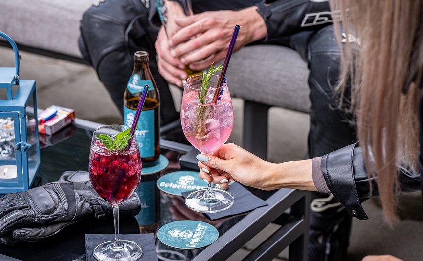 DAS UNTERRAIN lifestyle hotel **** © Armin Kleinlercher Zwei verschiedene Cocktails und eine Bierflasche auf einem Tisch mit Personen im Hintergrund