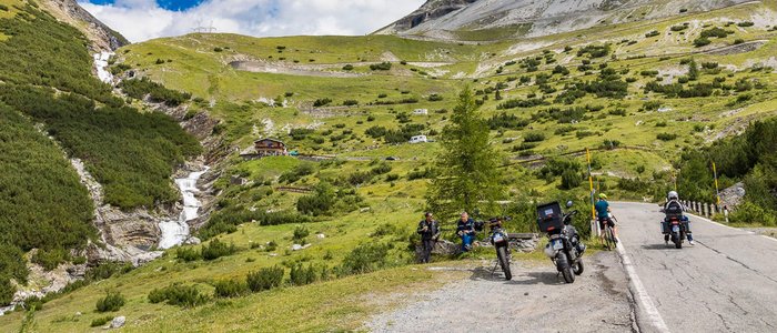 Bergstraße mit Motorradfahrern, Wanderern und Wasserfall in den Alpen
