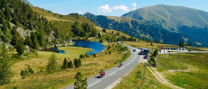 Gebirgsstraße mit Motorradfahrern, See und bewaldeten Bergen im Sommer