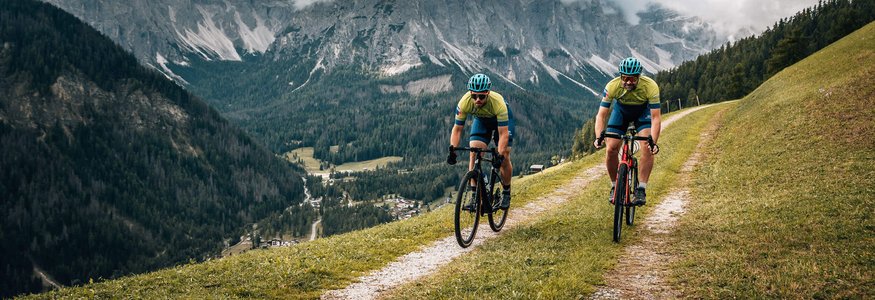 Gravel-Wochenende in San Vigilio © Jens Scheibe Zwei Radfahrer fahren auf einem Bergweg mit Alpen im Hintergrund