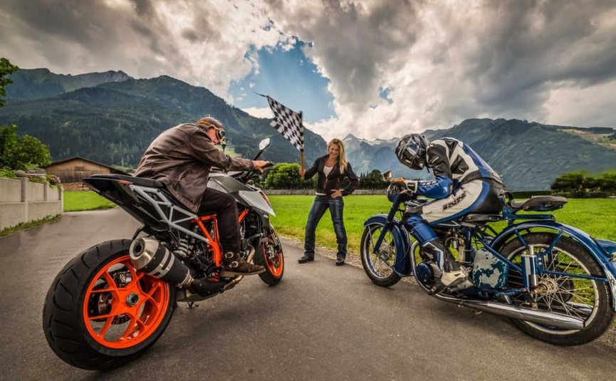Kurvenkönig Special ab € 208,- pro Person Zwei Motorradfahrer am Start mit Frau und Zielflagge vor Bergkulisse