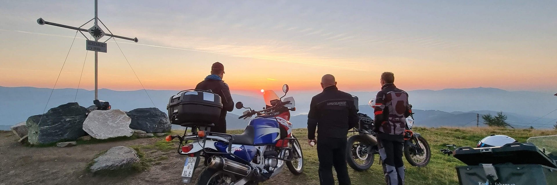 Drei Motorradfahrer schauen bei Sonnenuntergang vom Berggipfel mit Gipfelkreuz