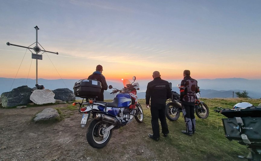 Motorradurlaub in der Ferienregion Wolfsberg – Region Lavanttal © Viktor Sator Drei Motorradfahrer schauen bei Sonnenuntergang vom Berggipfel mit Gipfelkreuz