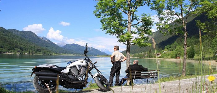 Motorrad am See mit zwei Männern, Bergen und blauem Himmel im Hintergrund