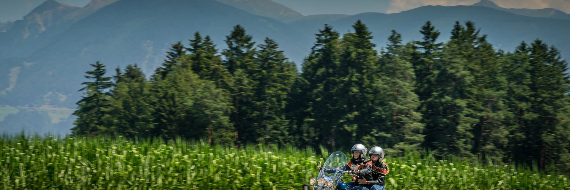 Zwei Motorradfahrer fahren auf Landstraße vor Wald und Bergen