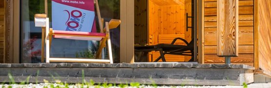 Liegestuhl auf Terrasse vor Holzhaus mit grünem Rasen im Vordergrund