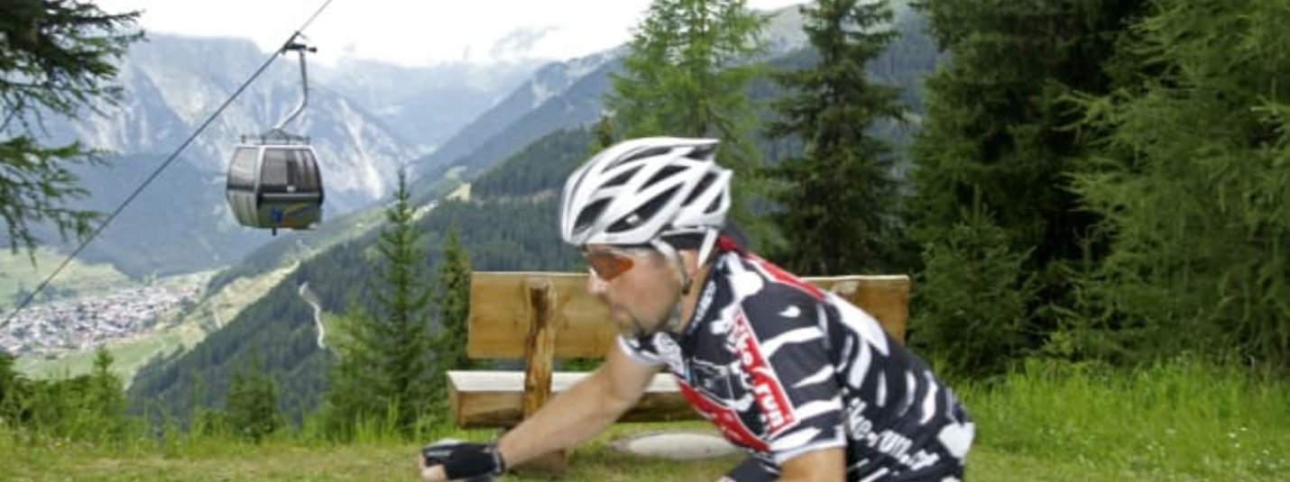 Mountainbikeurlaub Radrennfahrer fährt auf Bergweg mit Seilbahn und Wald im Hintergrund