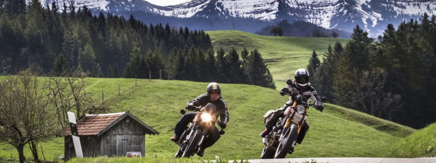 Kurvenwoche im Bergblick Zwei Motorradfahrer fahren auf Landstraße vor Alpen mit Gras und Wald