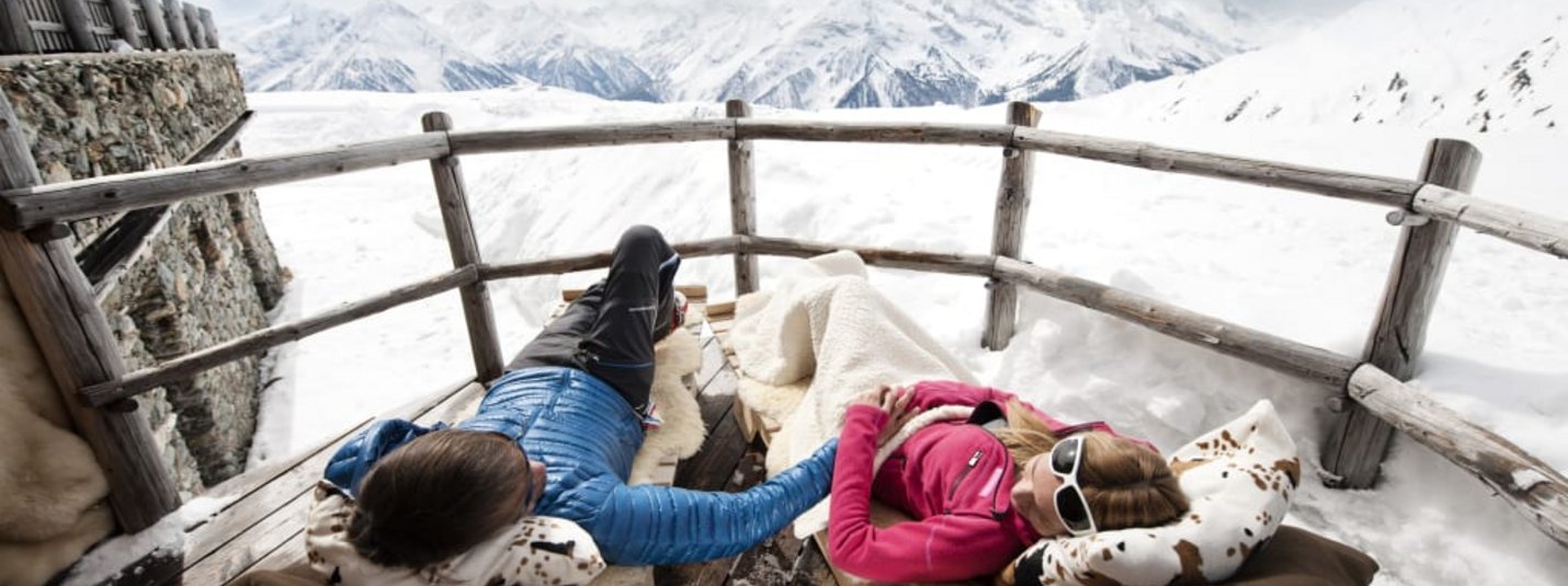Sonnenskilauf 4=3 Paar entspannt im Schnee auf Holzliegen mit Bergblick