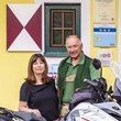Älteres Paar steht mit Motorrad vor gelbem Haus mit grünem Tor
