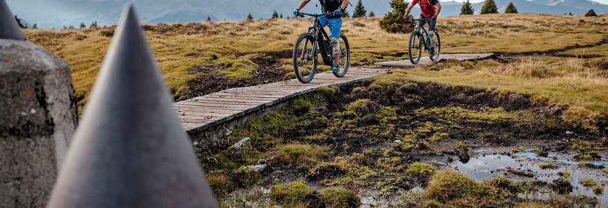Zwei Mountainbiker fahren auf einem Holzsteg in bergiger Landschaft
