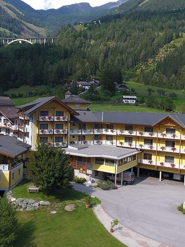 Motorradurlaub © Erlebnishotel Kärnten Mölltal Gelbes Hotelgebäude in grüner Berglandschaft mit Autobahnbrücke im Hintergrund