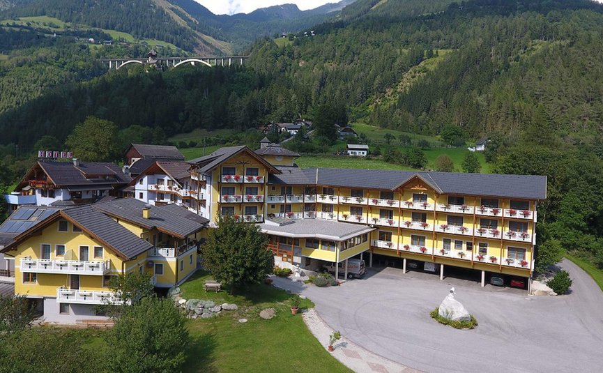 Erlebnishotel Kärnten-Mölltal ***S © Erlebnishotel Kärnten Mölltal Gelbes Hotelgebäude in grüner Berglandschaft mit Autobahnbrücke im Hintergrund