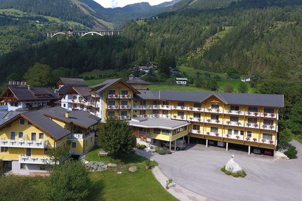 Erlebnishotel Kärnten-Mölltal © Erlebnishotel Kärnten Mölltal Gelbes Hotelgebäude in grüner Berglandschaft mit Autobahnbrücke im Hintergrund