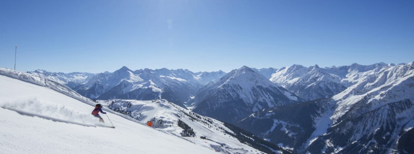 Sonnenskilauf 7=6 Zwei Skifahrer auf schneebedecktem Hang mit sonnigem Himmel und Alpen im Hintergrund