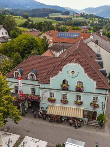 Altes Gasthaus mit blauen Fassade und Blumen in einem Dorf mit Bergen im Hintergrund